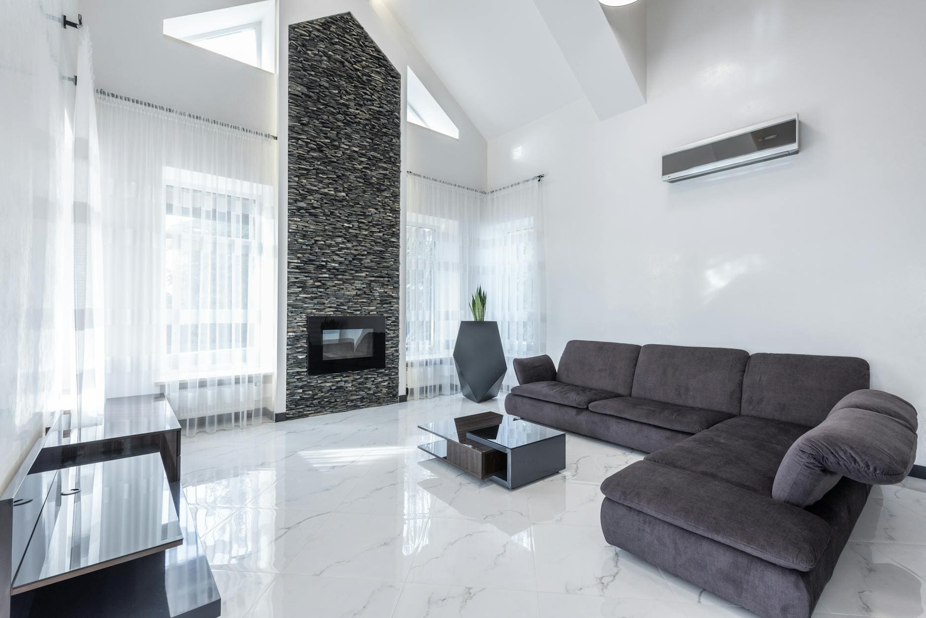 Helder moderne woonkamer met grijze bank, glazen salontafel, natuurstenen schouw en witte gordijnen.